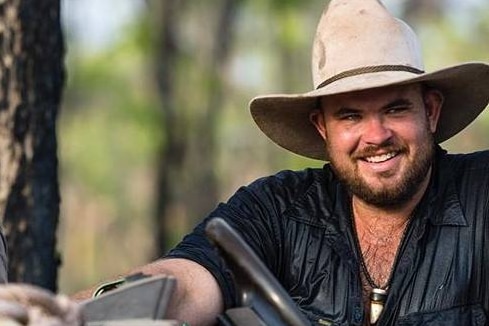 A smiling, bearded white man wearing a cowboy hat and a dark, wet, open-necked shirt.