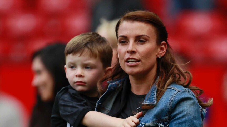 Coleen Rooney holds her son while standing on a football field in a denim jacket
