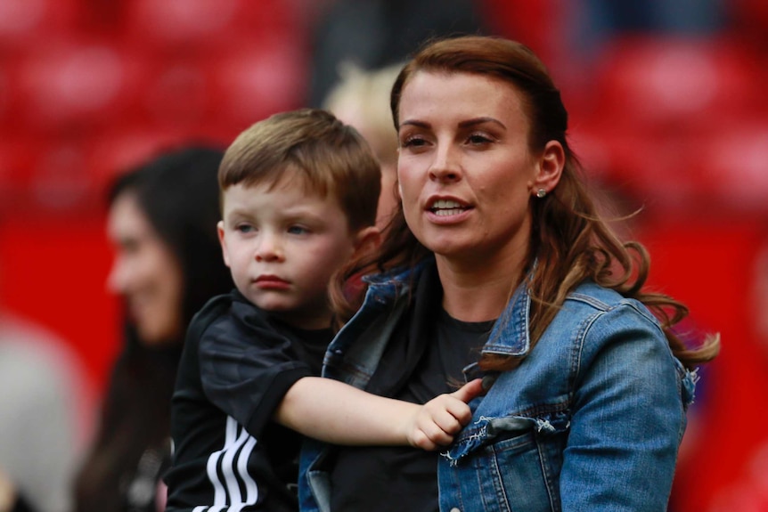 Coleen Rooney holds her son while standing on a football field in a denim jacket