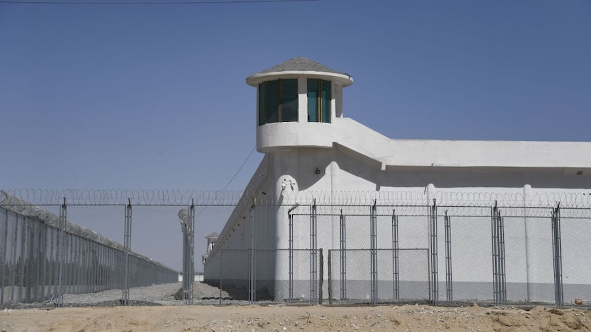white building with watchtower sits behind security fence with barbed wire in front of dirt