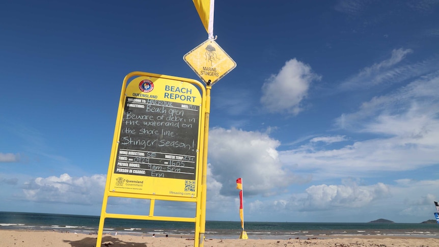 A chalk board stands on a beach warning beach goers of the conditions as people gather further down at the water's edge.