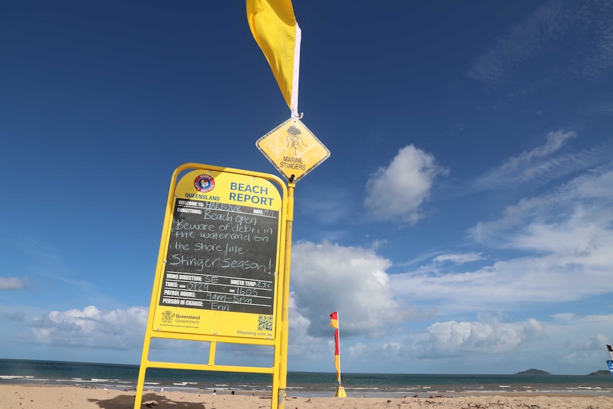 A chalk board stands on a beach warning beach goers of the conditions as people gather further down at the water's edge.