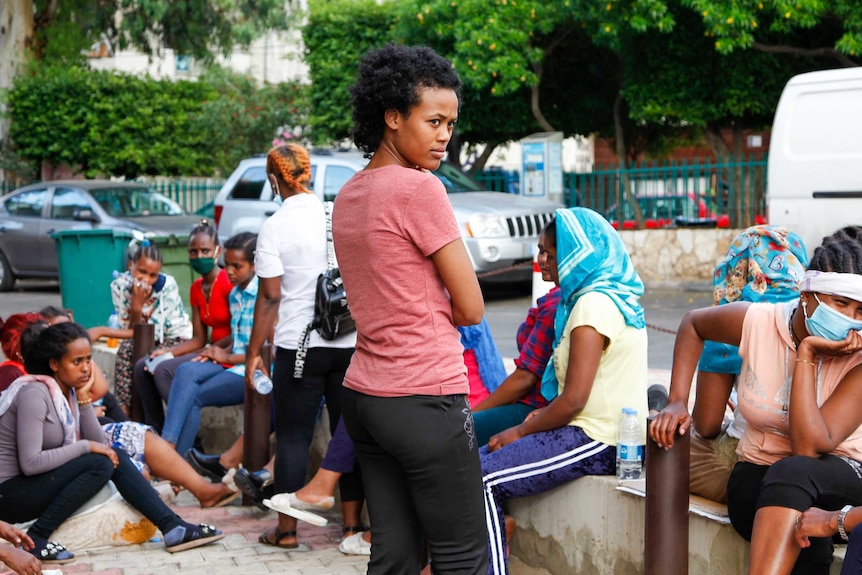 A young woman stands looking off in the distance surrounded by women sitting on a footpath