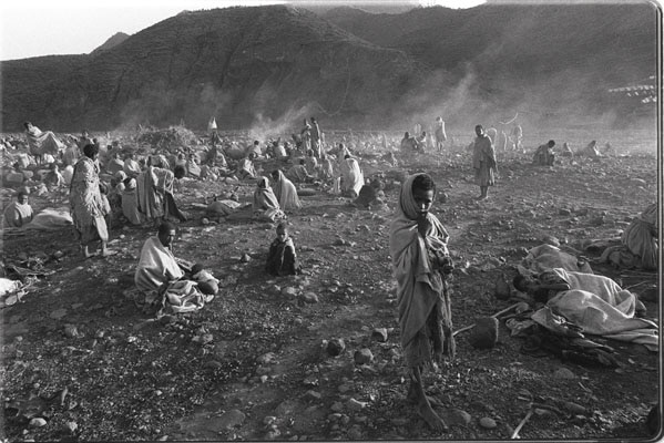 Emaciated children and adults stand and sit in a desolate landscape