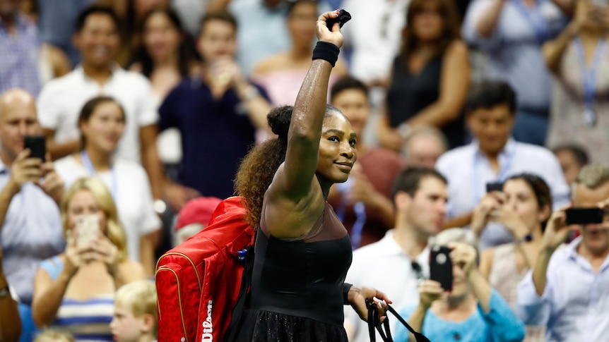 Serena Williams holds her hand up to acknowledge the crowd at the US Open as she carries her tennis bag off court.
