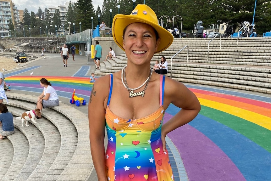 A trans woman wearing a rainbow dress and a yellow hat, stands on a rainbow walkway.