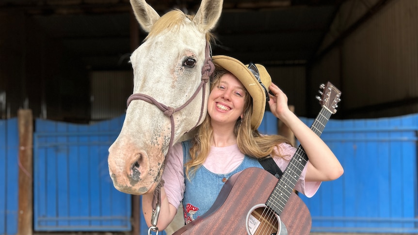 A woman holding a guitar and smiling next to a horse