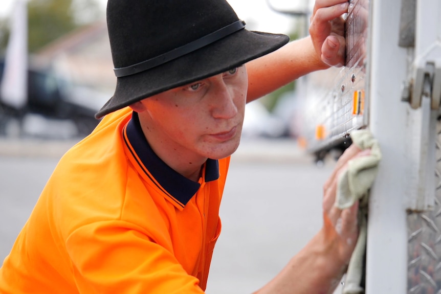 Nathan in a high vis orange shirt, black hat, wiping down the side of a truck.