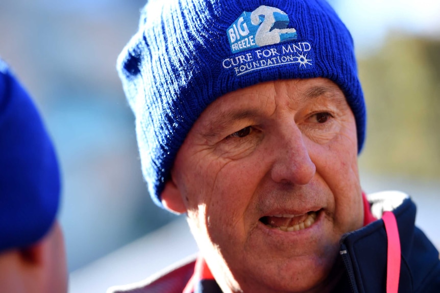 A middle-aged man in a thick beanie with the logo of a charity on it stands outside, speaking.