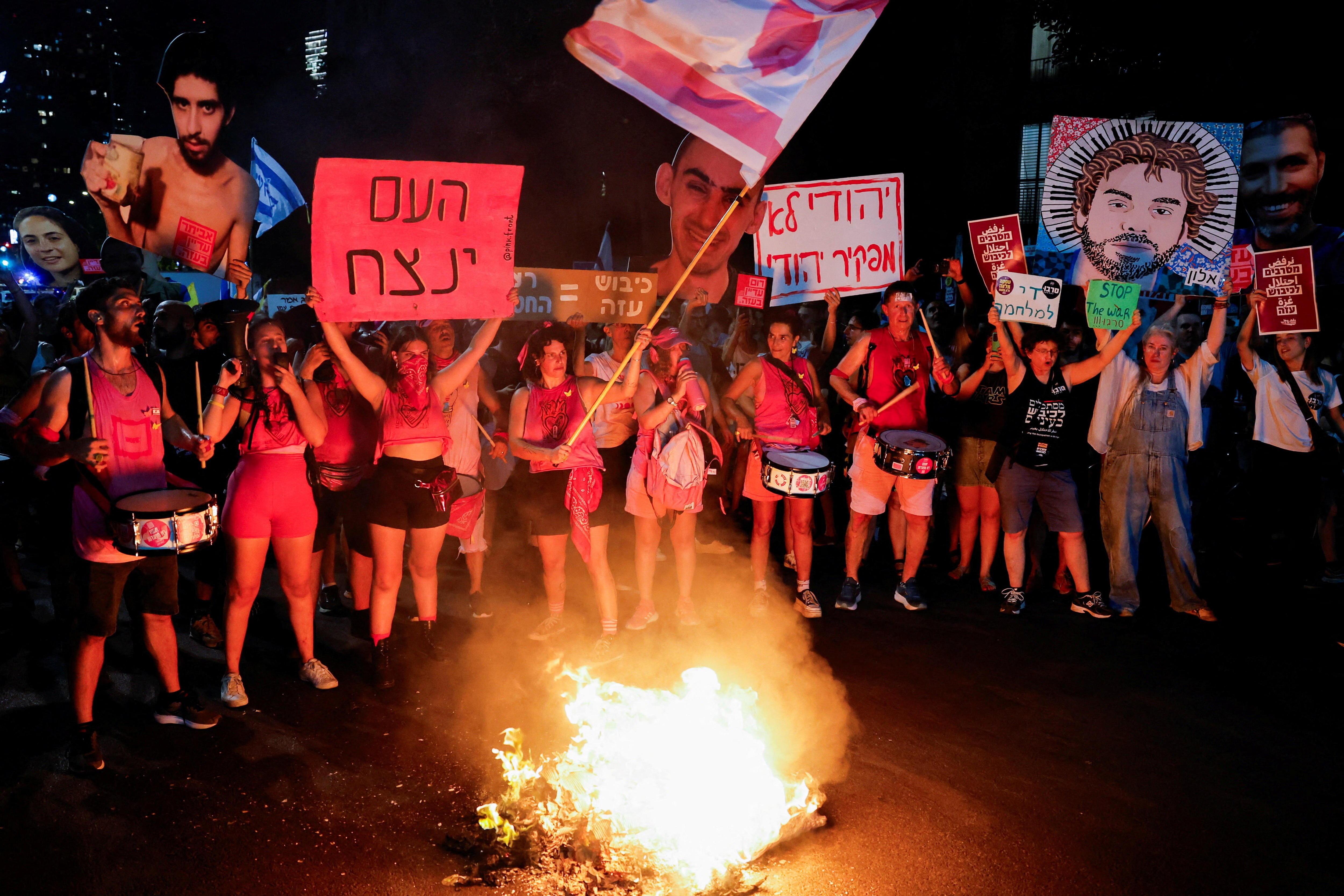 Protesters standing around a small fire wearing pink and holding up signs with the faces of Israeli hostages in Gaza