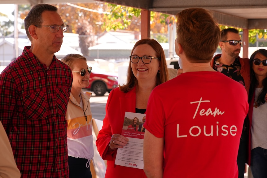 A woman wearing a red blazer speaks to a man wearing a red T-shirt with the words Team Louise on it