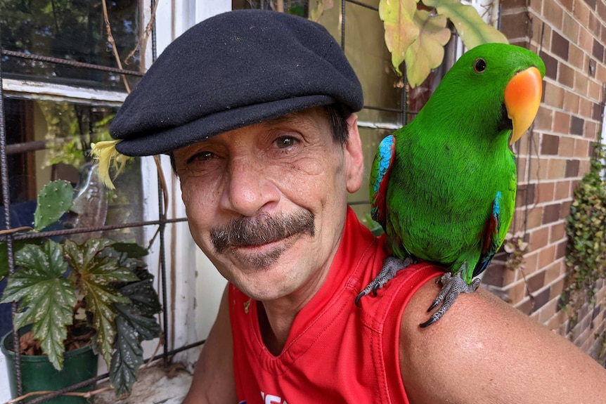 Peter "Pierre" Gawronski wearing a flatcap and with a bright green parrot on his shoulder, looks into the camera.