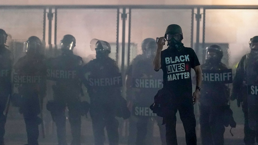 A protester wearing a Black Lives Matter t-shirt holds up a phone as he stands in front of riot police.