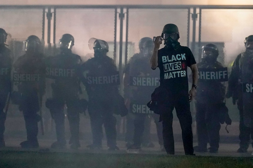 A protester wearing a Black Lives Matter t-shirt holds up a phone as he stands in front of riot police.