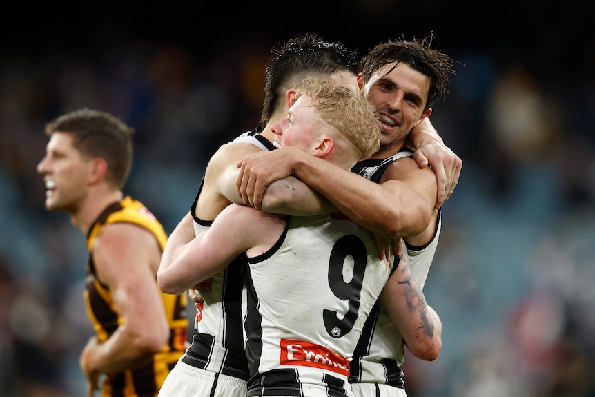 Three Collingwood players hug as Hawthorn players look disappointed behind them