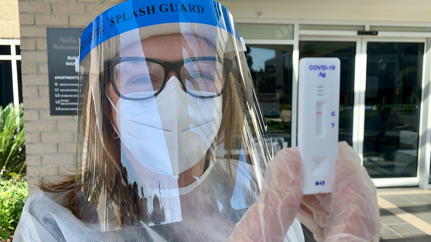 A woman wearing PPE, including face shield, holding a rapid antigen test.