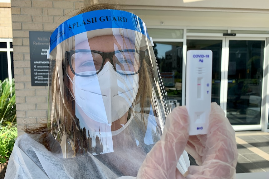 A woman wearing PPE, including face shield, holding a rapid antigen test.