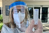 A woman wearing PPE, including face shield, holding a rapid antigen test.