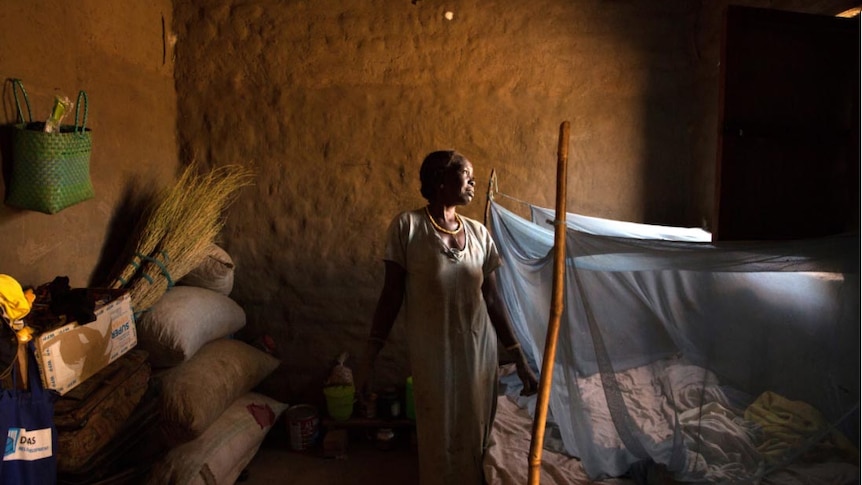 A south sudanese woman stands in front of a mud brick wall and looks out towards the window. 