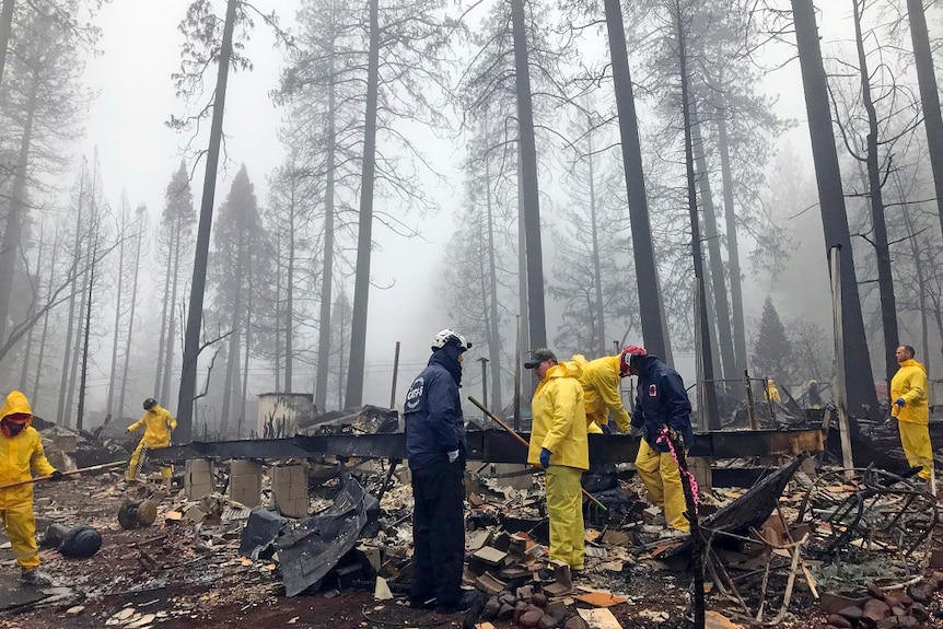 Seven adults in protective gear use tools as they search through charred wreckage looking for human remains.