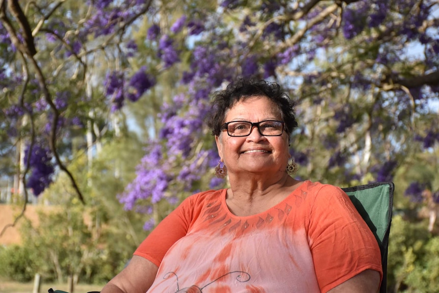 Aunty Brenda sits in a park with jacaranda trees behind.