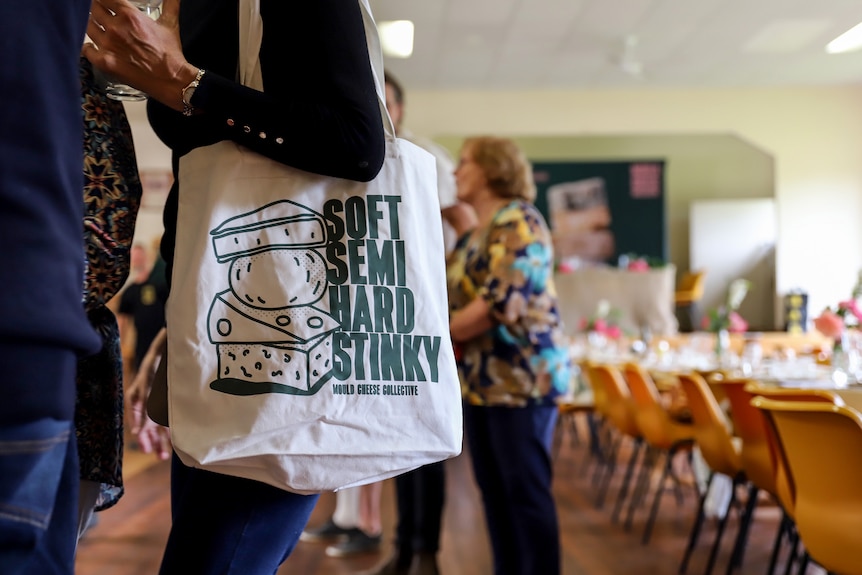 A tote bag hangs off a woman's shoulder with a hall and table in background. The bag reads Soft Semi Hard Stink