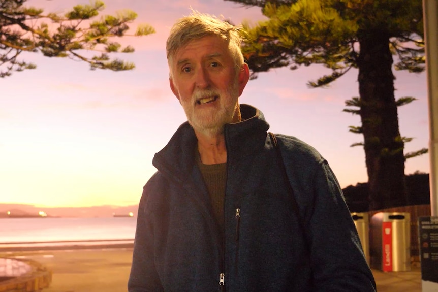 An older, bearded man with grey hair smiles on a beach lit by a low sun.