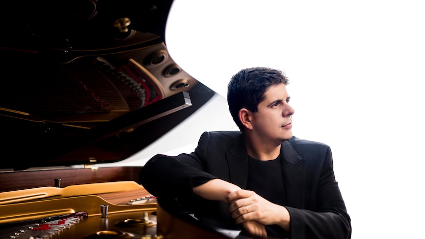 Pianist Javier Perianes rests his arm on the bow of the piano looking away. He is wearing a black tee shirt and a black jacket
