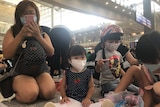 A woman kneels next to three children drawing pictures while sat on the ground at the airport. They all wear surgical masks.