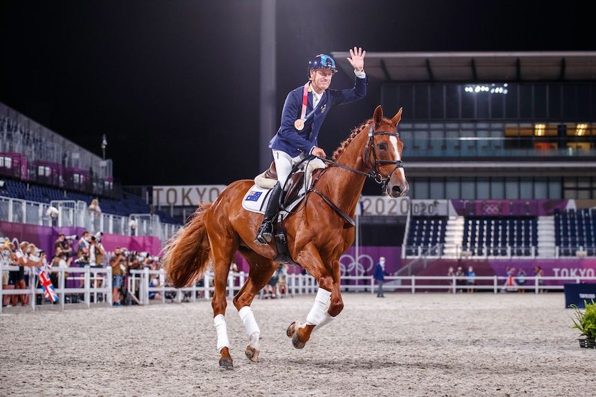 A man on a horse rides in an arena and waves while wearing a medal around his neck