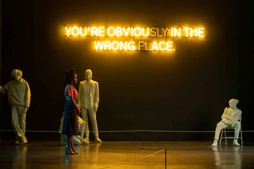 Teyonah Parris stands in a dark gallery illuminated by a yellow neon sign which reads "You're obviously in the wrong place".