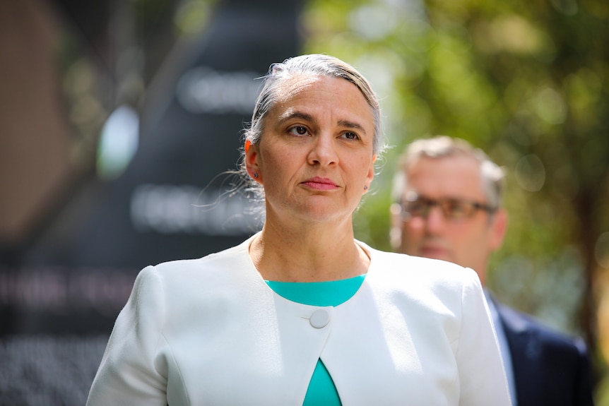 A woman dressed in a white jacket stands outside looking serious. 