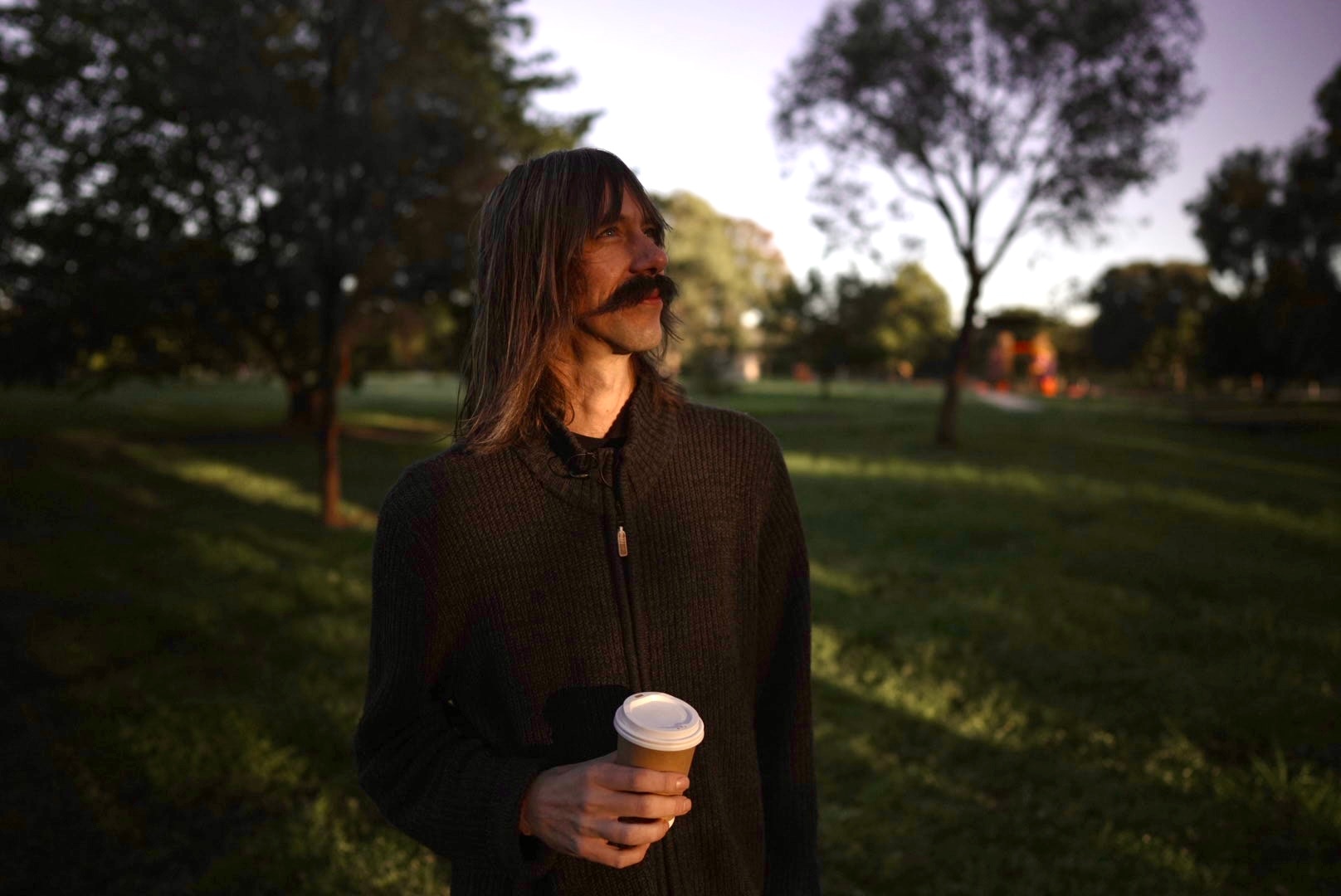 A man with a moustache holds a takeaway coffee cup and looks up.