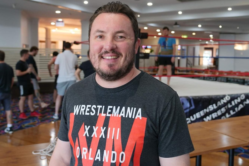 A man with brown hair and a bear wearing a wrestling themed T shirt smiles. He's standing in front of a wrestling ring.