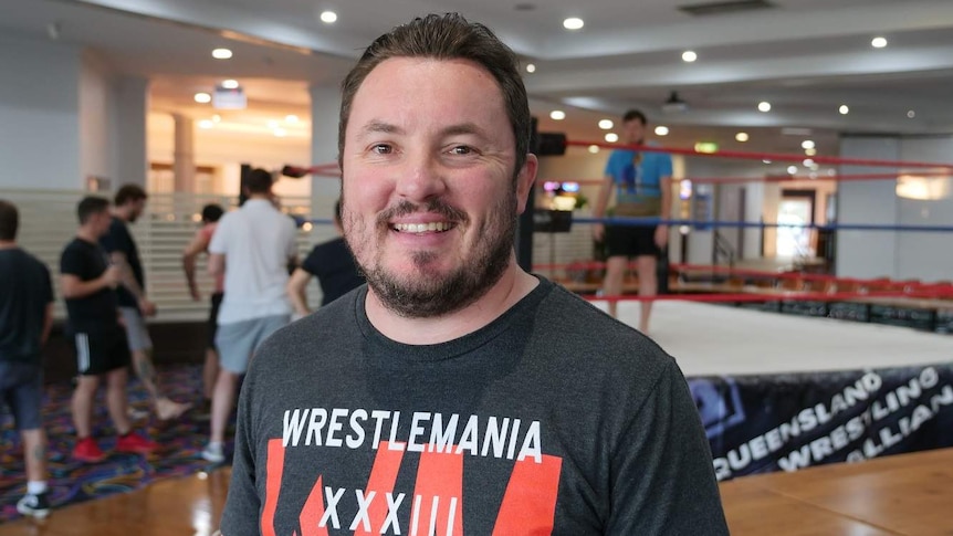 A man with brown hair and a bear wearing a wrestling themed T shirt smiles. He's standing in front of a wrestling ring.