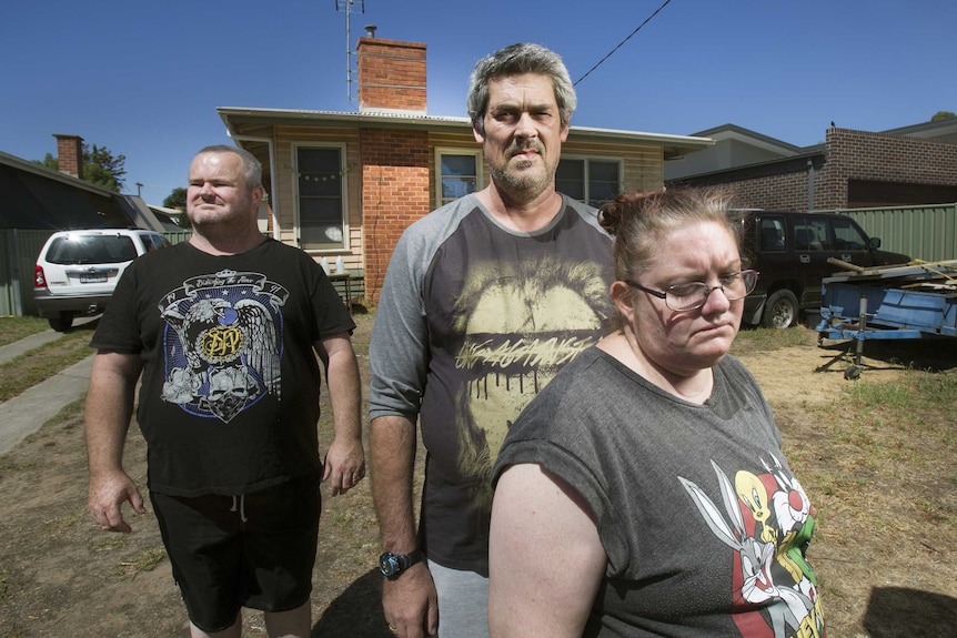 Peter Arthur, Ronald Lyons and Christine Lyons stand in the front yard of a suburban house.