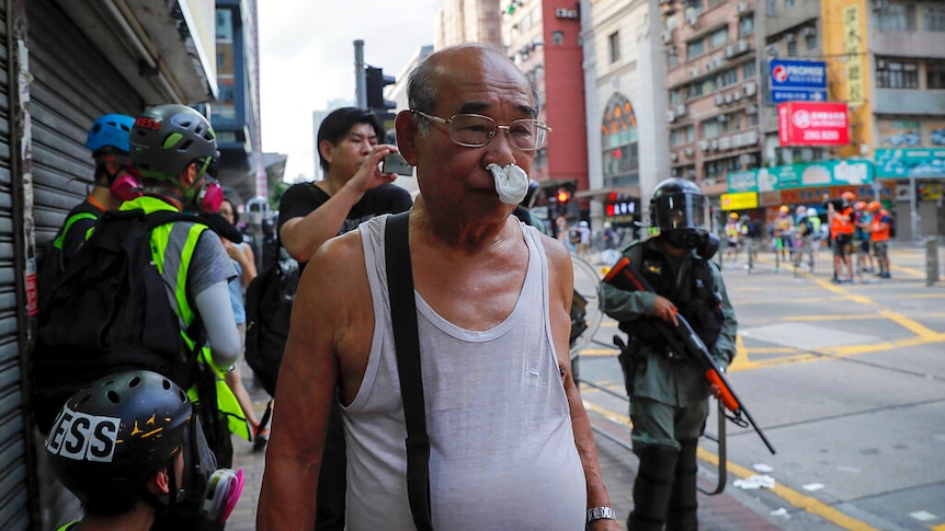 An elderly man wearing glasses and a white singlet has tissues blocking his nose as he reacts from tear gas being released.