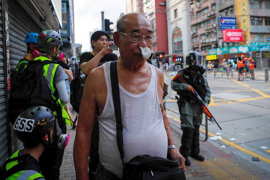 An elderly man wearing glasses and a white singlet has tissues blocking his nose as he reacts from tear gas being released.