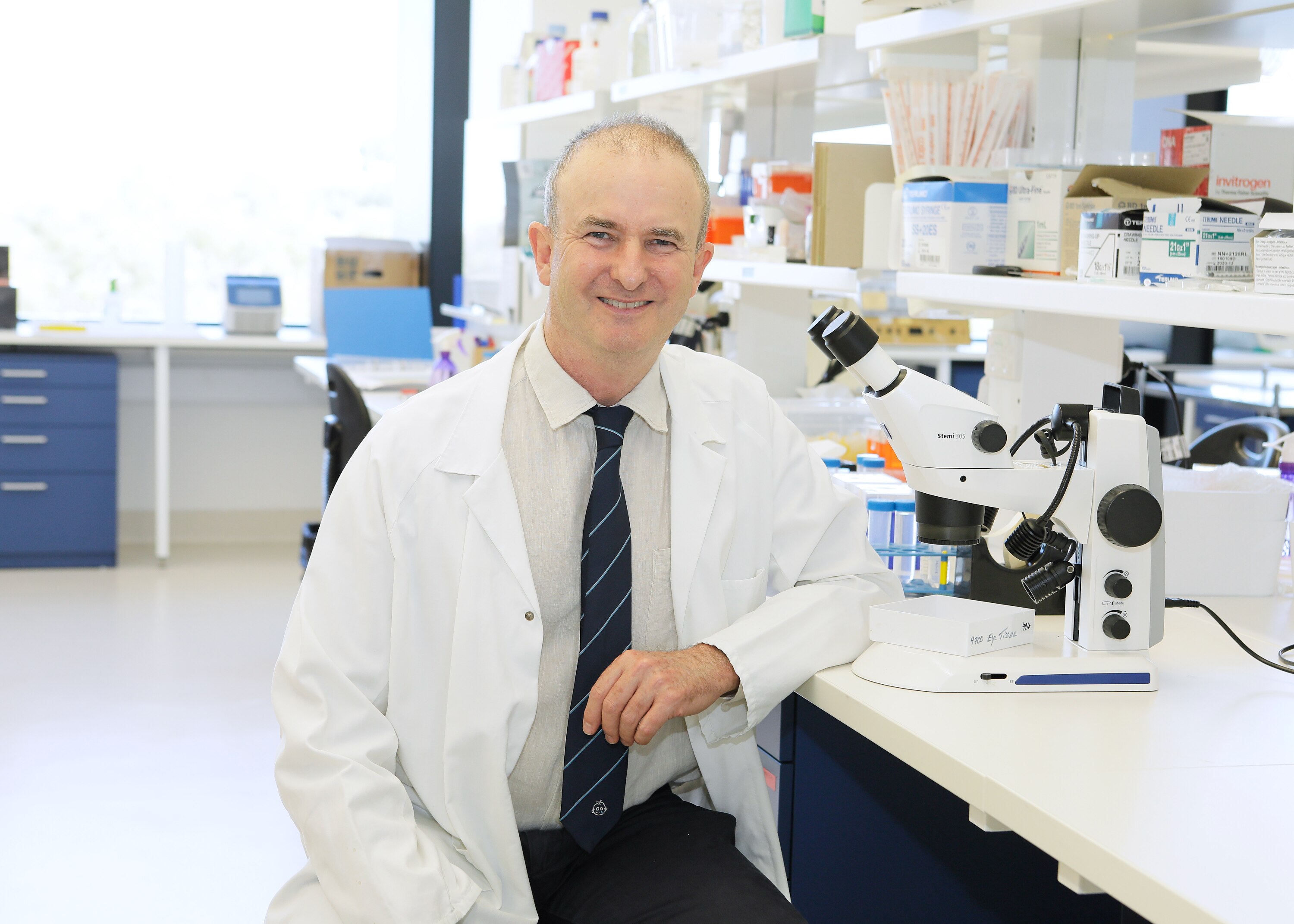 Smiling man in a lab coat sits at a desk next to a microscope.