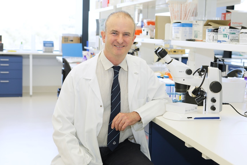 Smiling man in a lab coat sits at a desk next to a microscope.