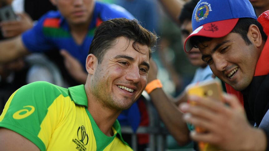 Marcus Stoinis smiles while leaning towards a fan wearing an Afghanistan Cricket Board cap, who is holding a phone