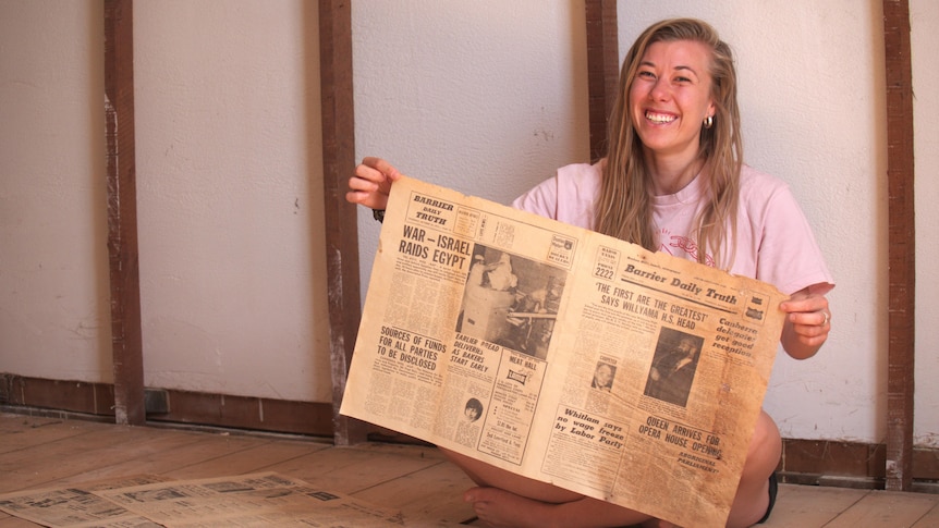A woman with blonde hair smiling while sitting down and holding newspapers