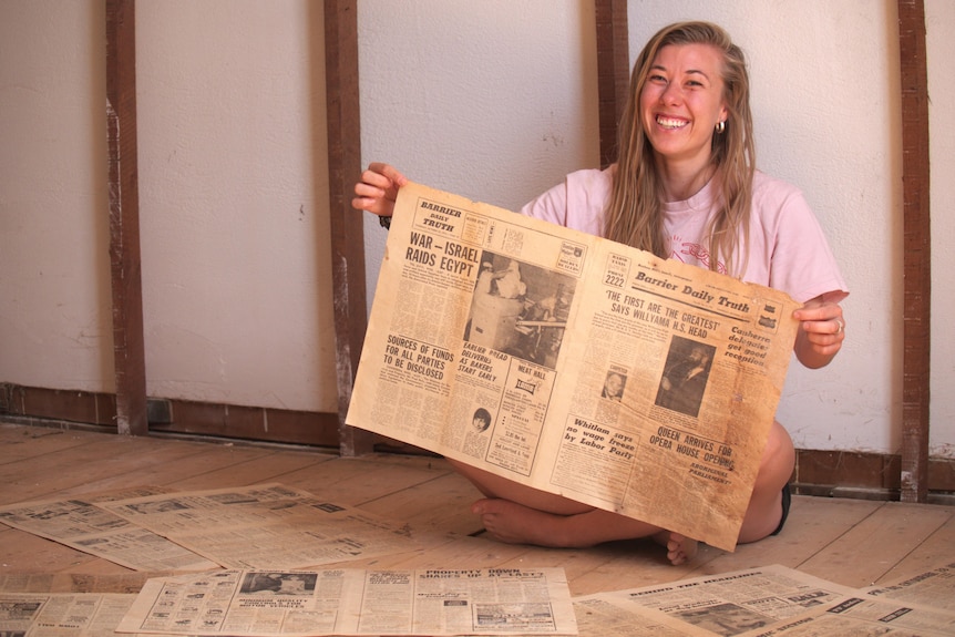 A woman with blonde hair smiling while sitting down and holding newspapers