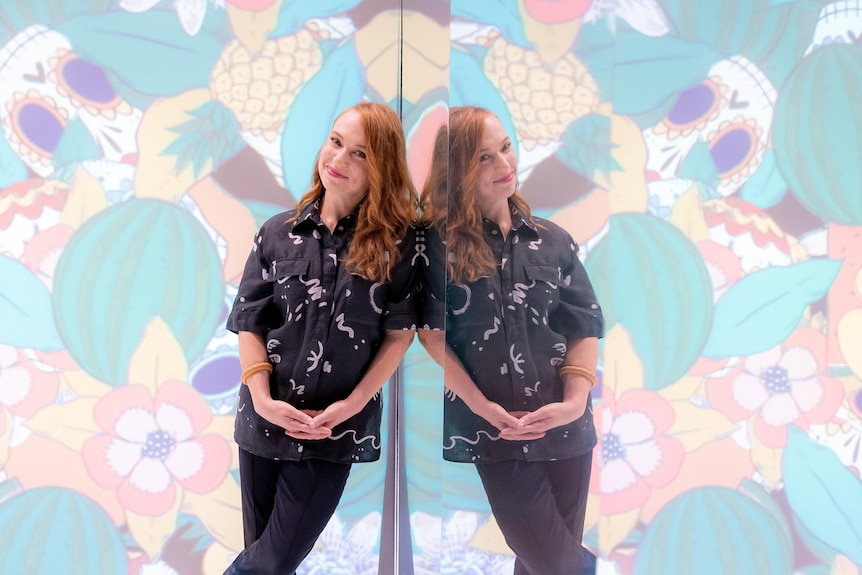 A red-haired white woman in a black shirt with white shapes and black pants leans against mirror in a colourful gallery