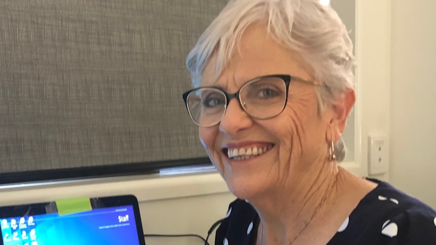 An older, bespectacled woman smiles warmly while sitting at a work desk.