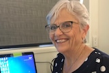 An older, bespectacled woman smiles warmly while sitting at a work desk.