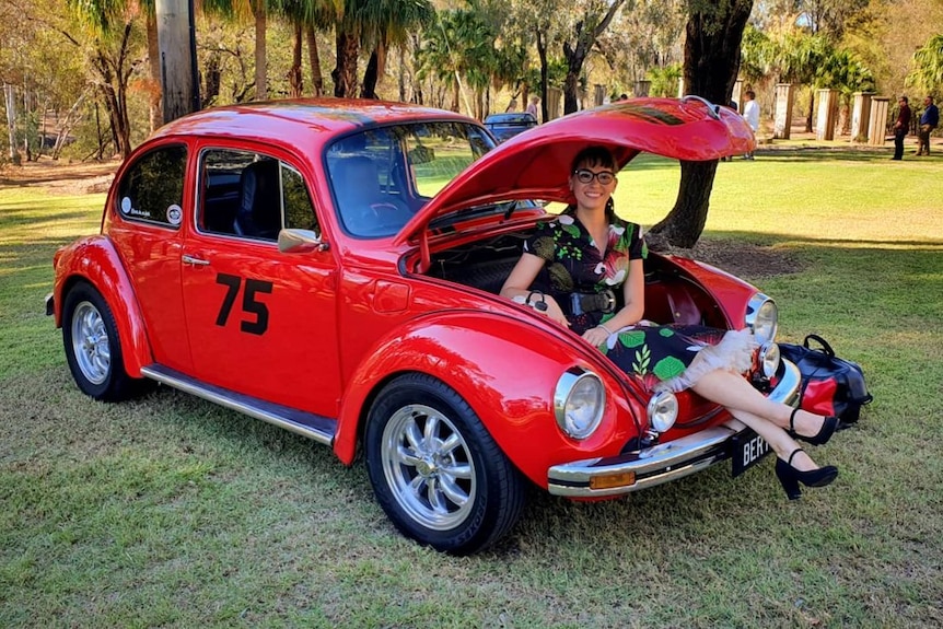 A young woman in a dress and heels, sitting in a VW Beetle bonnet, smiling.