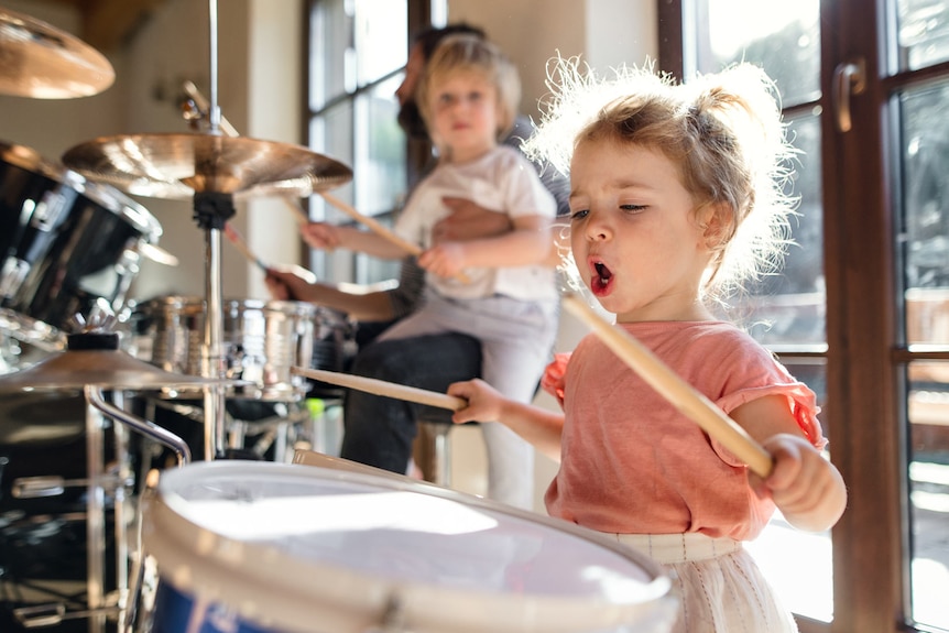A very young girl is beating on a drum with drumsticks. Her mouth is open as she sings along.