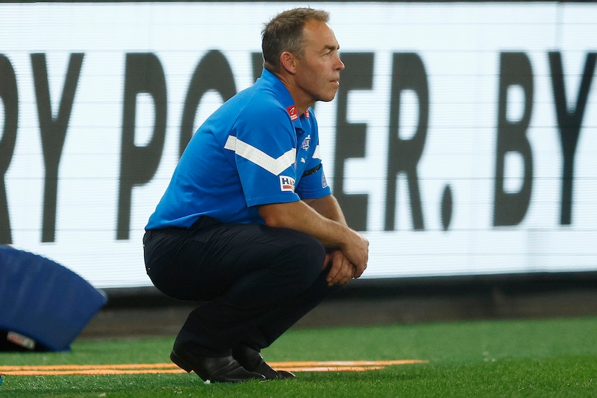 Alastair Clarkson crouching as he watches the Kangaroos play the Eagles in the AFL.