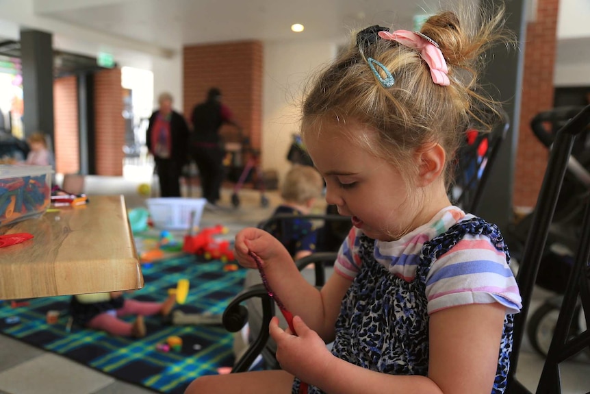 Young girl with her hair tied back in a pink bow makes a necklace out of painted pasta at a table.